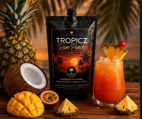 A stand-up pouch of Tropicz Rum Punch cocktail mix by Gold Coast stands on a wooden surface, surrounded by tropical ingredients including a whole pineapple, a cut coconut, mango pieces, pineapple slices, a passion fruit half, and a glass of orange-red punch garnished with pineapple and a cherry. The background shows blurred palm leaves and warm sunset tones.